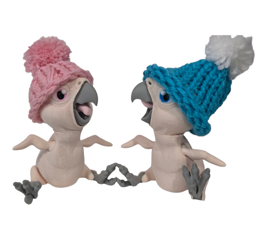 Two 3D-printed articulated bald parrots wearing hand-knitted blue and pink pom-pom beanies, posed playfully together.